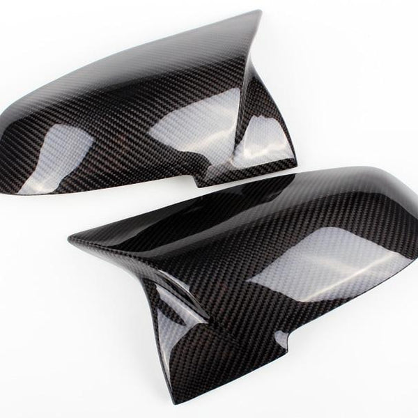 Two glossy carbon fiber mirror covers are positioned face-up on a plain white background, showcasing their sleek, aerodynamic design and textured pattern.