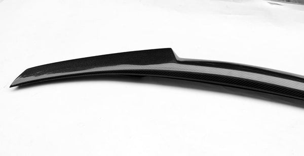 A sleek carbon fiber car spoiler, featuring a glossy finish, rests on a plain white background, highlighting its aerodynamic curve and textured surface.