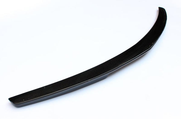 A curved, black carbon fiber car spoiler rests on a white surface, showcasing its sleek, aerodynamic design.