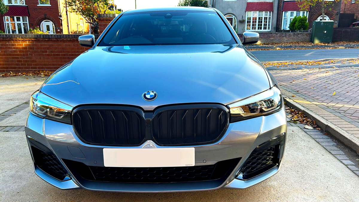A silver BMW car with a prominent black grille is parked on a driveway. Surrounding it are brick houses and a street lined with autumn leaves.