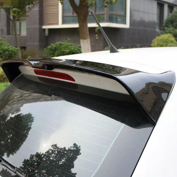 A car rear end with a spoiler and antenna is parked outdoors near a building, surrounded by greenery and trees. Reflections are visible on the rear window.