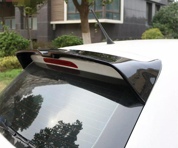 A car rear end with a spoiler and antenna is parked outdoors near a building, surrounded by greenery and trees. Reflections are visible on the rear window.