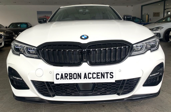 A white BMW car displays "CARBON ACCENTS" on the front license plate. The vehicle is parked indoors, surrounded by other cars, with visible headlights and kidney grille design.