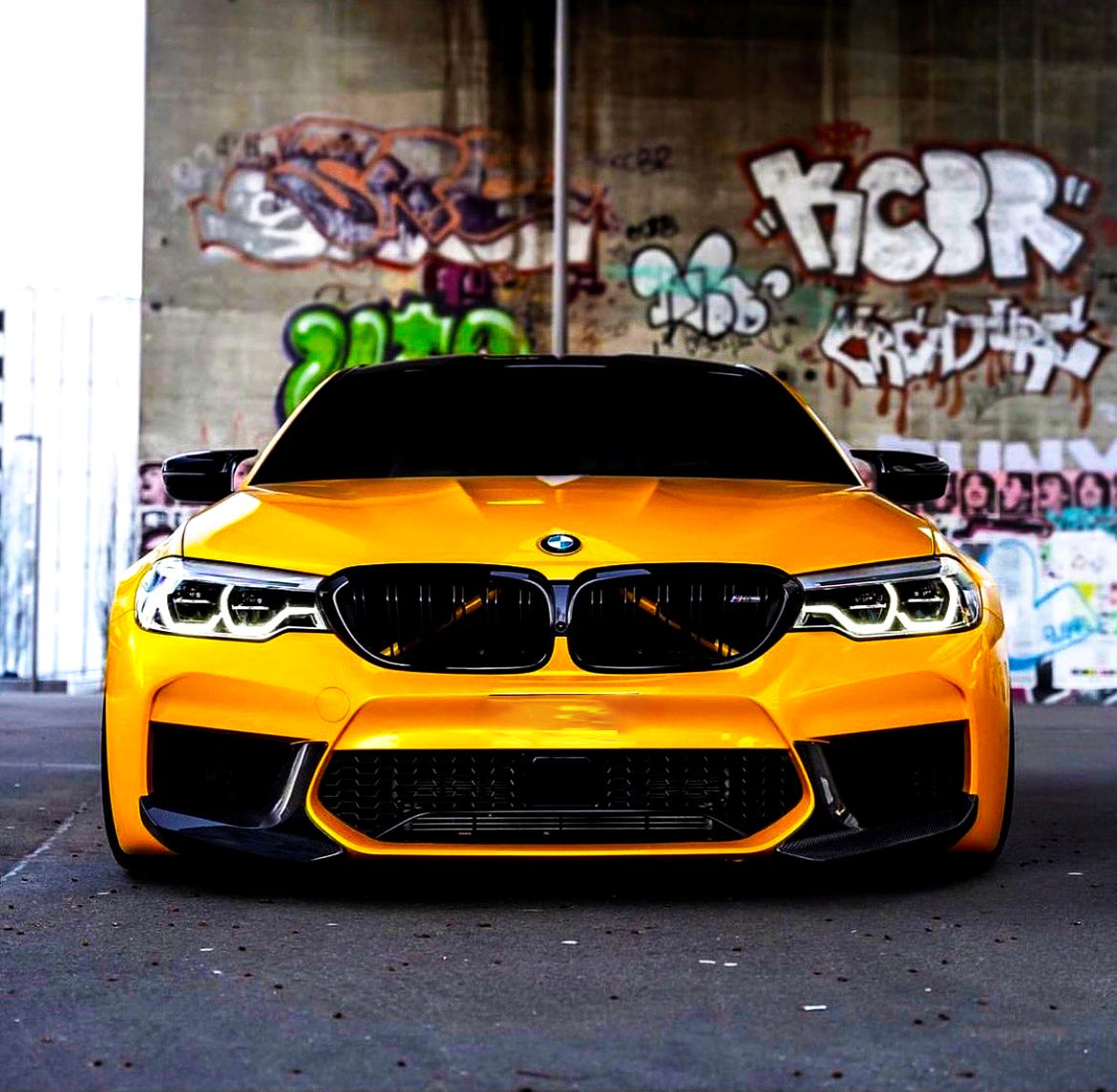 A bright yellow sports car, parked stationary, faces forward. It is under a graffiti-covered bridge, with colorful urban artwork like "HCER" visible on the concrete walls behind it.