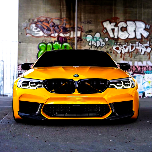 A bright yellow sports car, parked stationary, faces forward. It is under a graffiti-covered bridge, with colorful urban artwork like "HCER" visible on the concrete walls behind it.