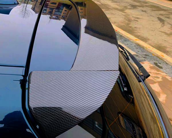 A carbon fiber car spoiler is mounted on a sleek black vehicle, reflecting nearby buildings and parked along a street with another car in the background.