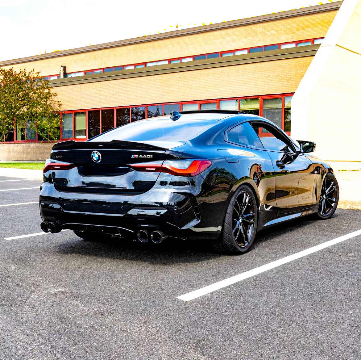 A sleek black BMW M440i is parked diagonally in a nearly empty parking lot, with a brick building featuring red-framed windows in the background.