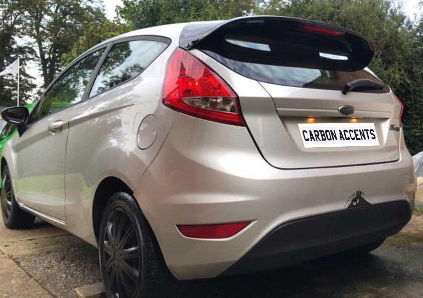 A silver car with tinted windows and a rear spoiler is parked on a driveway surrounded by trees. The license plate reads "CARBON ACCENTS."