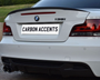 The car's rear features a white BMW 135i, with carbon fiber accents and "CARBON ACCENTS" license plate text. It is parked on gravel, with a wooden fence and grass in the background.