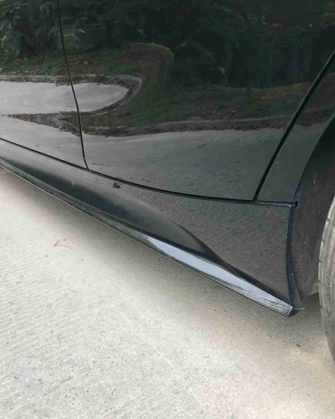 Car side skirts run along the bottom edge, enhancing aerodynamics, with black wheels on smooth pavement. Greenery is visible in the background.