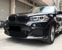 A black BMW SUV is parked on a concrete surface, its headlights and sleek grille visible. Nearby, an urban setting includes closed shutters and a bicycle.