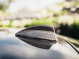 Shark fin antenna, made of carbon fiber, sits on a car's roof. It's in a sunlit outdoor environment with blurred greenery in the background.