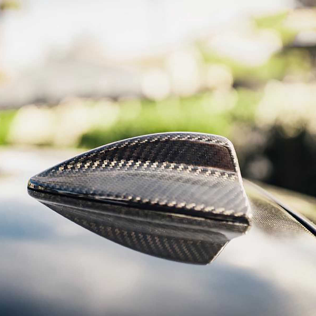 Shark fin antenna, made of carbon fiber, sits on a car's roof. It's in a sunlit outdoor environment with blurred greenery in the background.