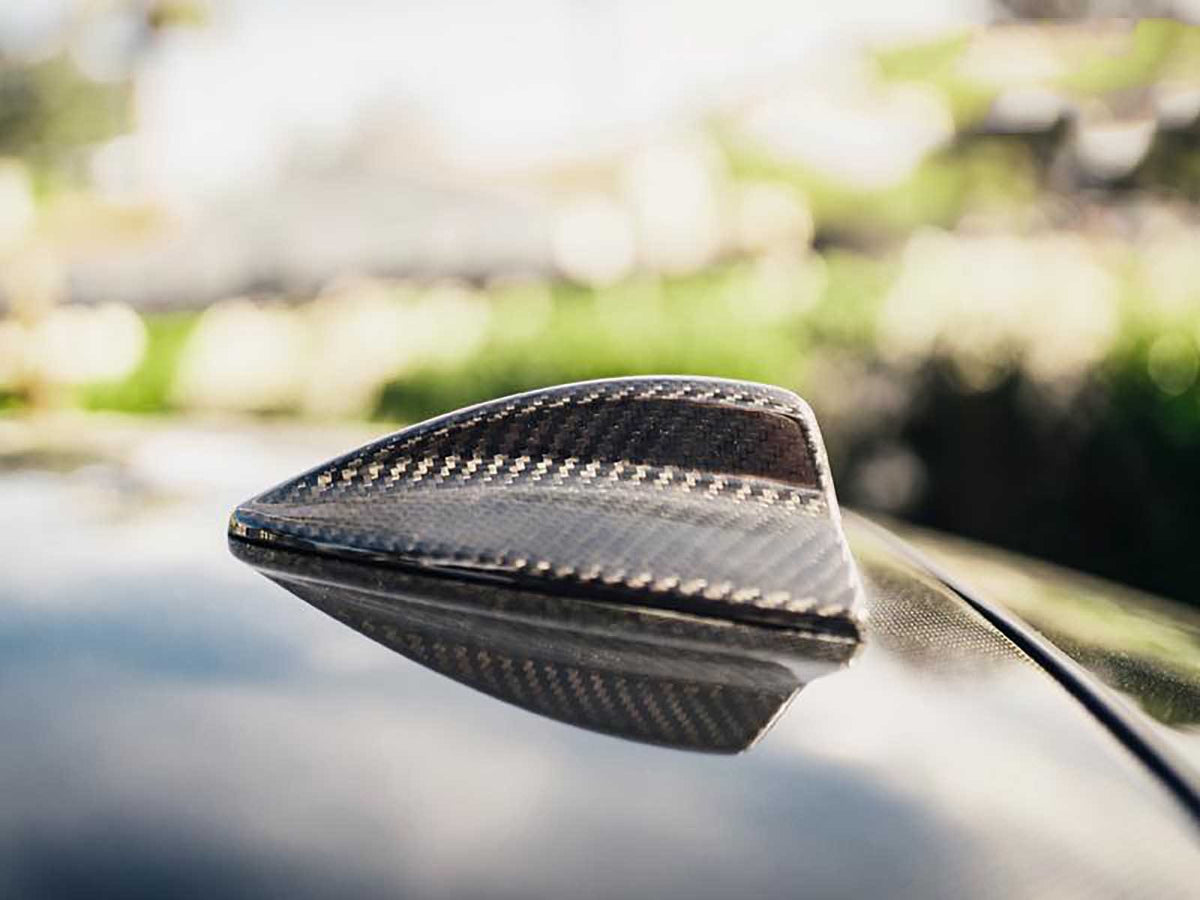 Shark fin antenna, made of carbon fiber, sits on a car's roof. It's in a sunlit outdoor environment with blurred greenery in the background.