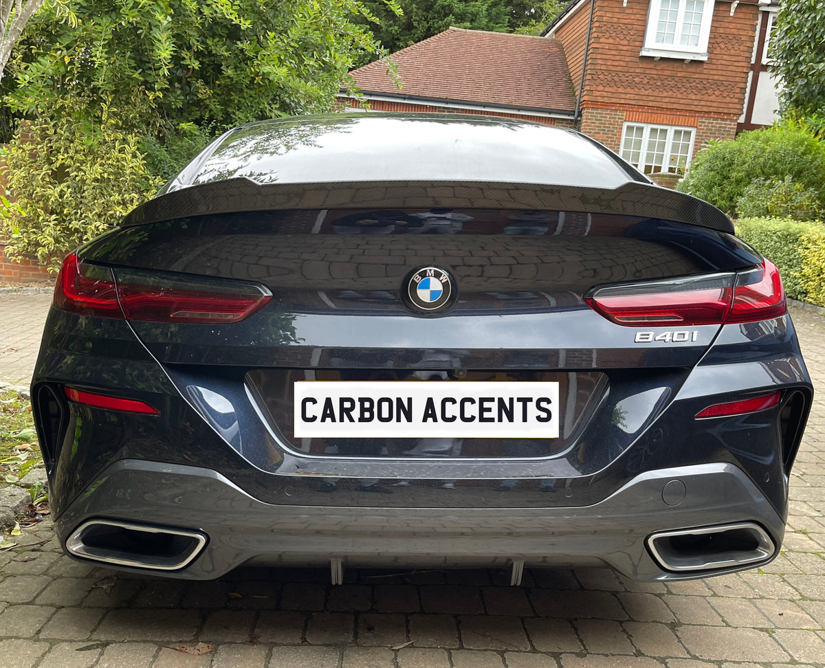 The car, a BMW 840i, is stationary with a license plate reading "CARBON ACCENTS," parked in a driveway surrounded by green bushes and a brick house in the background.