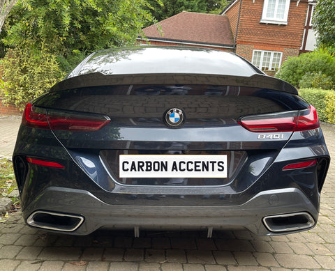 The car, a BMW 840i, is stationary with a license plate reading "CARBON ACCENTS," parked in a driveway surrounded by green bushes and a brick house in the background.