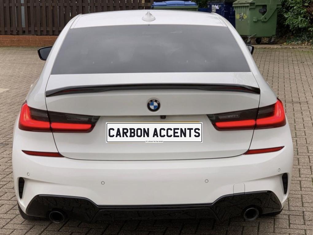 A white BMW parked on a paved surface, featuring rear carbon accents. The context includes a wooden fence and two trash bins. License plate reads: "CARBON ACCENTS.”