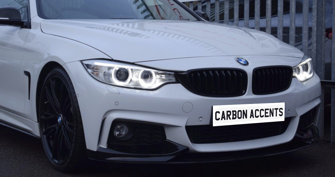 A white BMW car with illuminated headlights and black wheels is parked beside a metal fence. The license plate reads "CARBON ACCENTS."