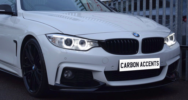 A white BMW car with illuminated headlights and black wheels is parked beside a metal fence. The license plate reads "CARBON ACCENTS."