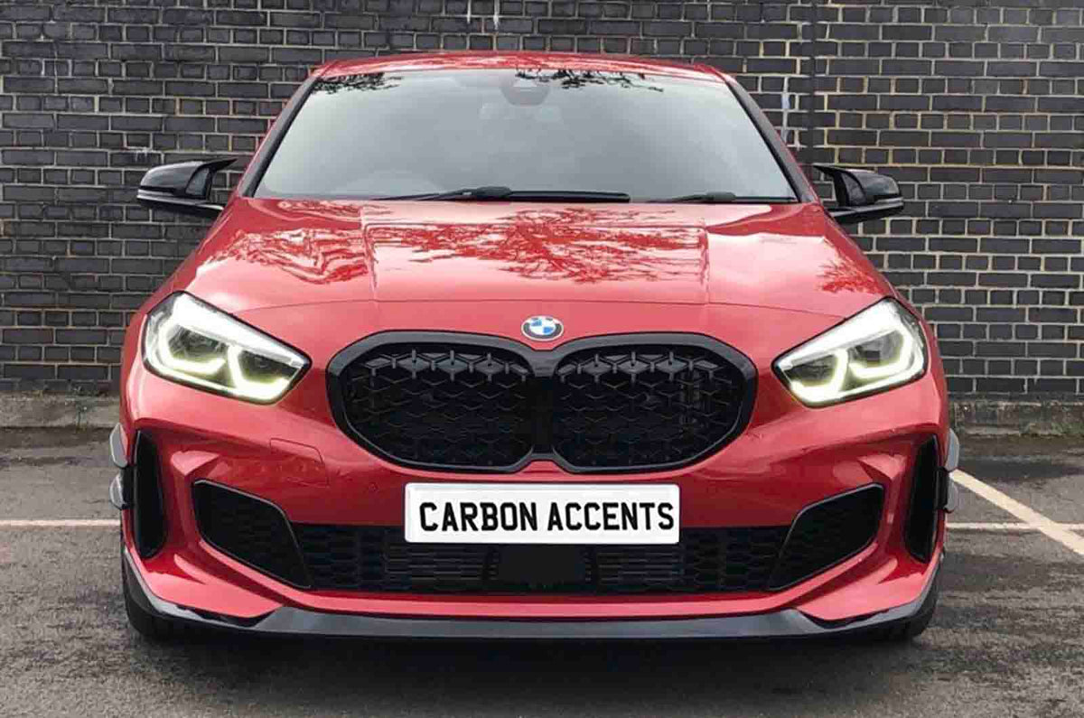 Red car with illuminated headlights is parked on asphalt, facing forward. It has a prominent black grille. The license plate reads "CARBON ACCENTS." A brick wall forms the background.