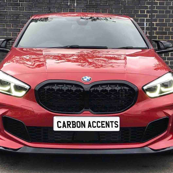 Red car with illuminated headlights is parked on asphalt, facing forward. It has a prominent black grille. The license plate reads "CARBON ACCENTS." A brick wall forms the background.