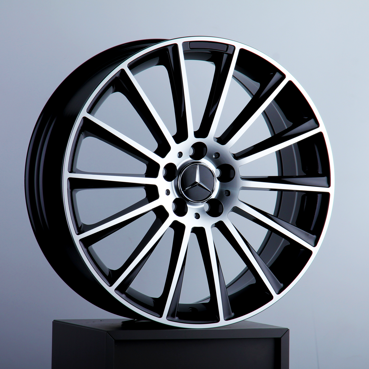 A silver and black alloy car wheel stands upright on a black pedestal. The central hub features the Mercedes-Benz logo. The background is a plain, light gray surface.