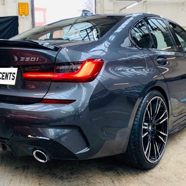 Car parked indoors, featuring a sleek gray BMW 320i with visible dual exhausts and tinted windows. The license plate reads "CARBON ACCENTS." The setting is an industrial garage.
