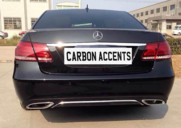 A black Mercedes-Benz car is parked on a concrete surface, displaying dual exhausts. The license plate reads "CARBON ACCENTS." Industrial buildings are visible in the background.