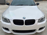 A white BMW car is parked on a concrete lot, facing forward, near a building with roll-up doors.