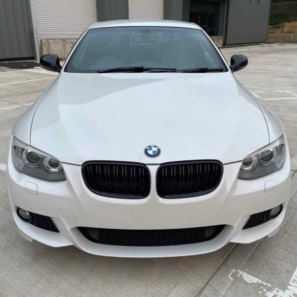 A white BMW car is parked on a concrete lot, facing forward, near a building with roll-up doors.