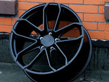 A black alloy wheel stands upright against an orange brick wall on a gray concrete surface. The wheel features multiple spokes and a central hub surrounded by five lug holes.