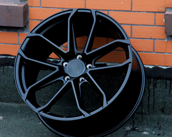 A black alloy wheel stands upright against an orange brick wall on a gray concrete surface. The wheel features multiple spokes and a central hub surrounded by five lug holes.