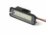 A rectangular LED light with a black housing and clear, textured lens is illuminated, featuring yellow diodes. It has red and black wires extending from the back, set against a plain background.