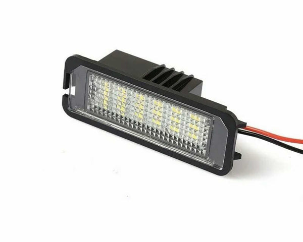 A rectangular LED light with a black housing and clear, textured lens is illuminated, featuring yellow diodes. It has red and black wires extending from the back, set against a plain background.