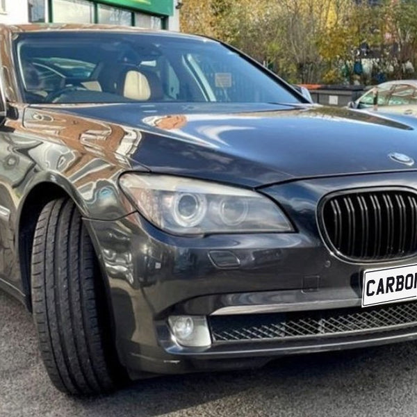 A sleek, dark-colored car with the front wheel turned slightly to the side is parked on a street. The license plate reads "CARBON ACCENTS," surrounded by other parked vehicles.