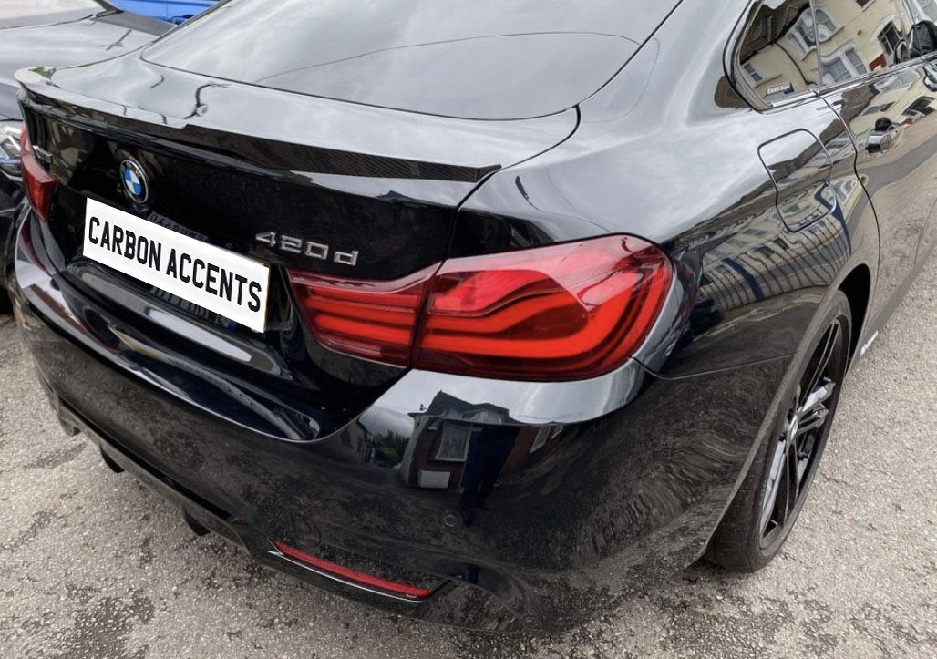 Car with license plate reading "CARBON ACCENTS" is parked. It's a black BMW 420d, featuring red taillights, set on a paved area with buildings reflected on its surface.