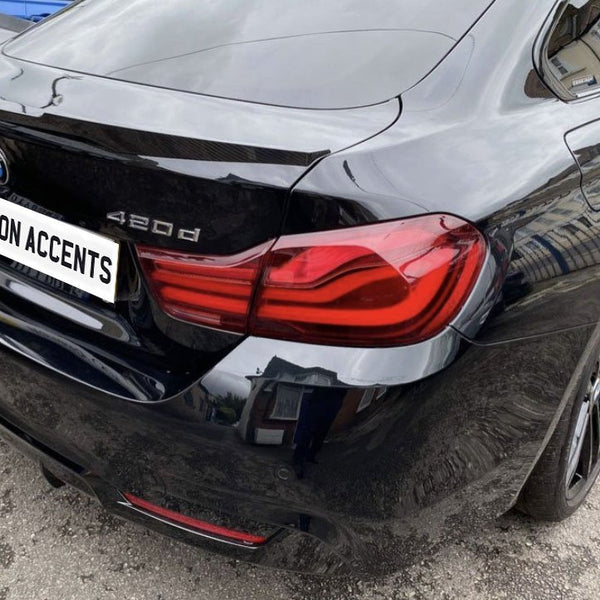 Car with license plate reading "CARBON ACCENTS" is parked. It's a black BMW 420d, featuring red taillights, set on a paved area with buildings reflected on its surface.