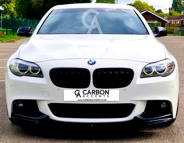 A white BMW sedan is parked, facing forward, under clear skies in a parking lot with greenery and buildings. Text on the front reads: "CARBON ACCENTS www.CARBONACCENTS.CO.UK".