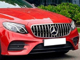 A red Mercedes-Benz car with a prominent grille and headlights is parked on a street, with lush green bushes and a glass building in the background.