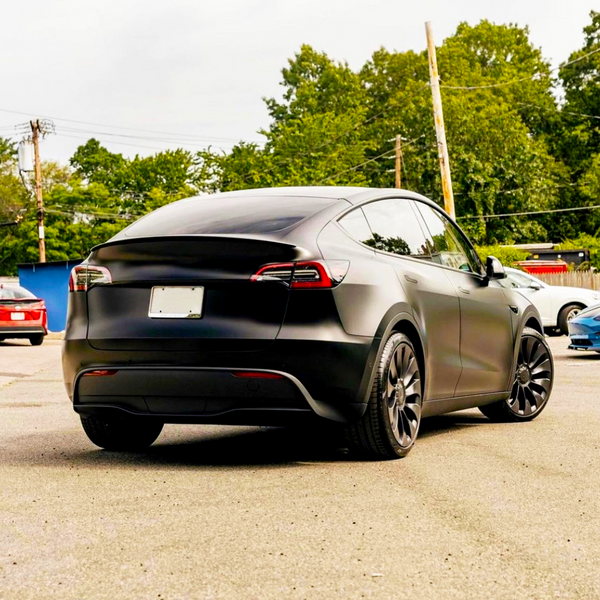 A matte black car is parked in a lot, showing a sleek rear view. In the background, other vehicles and greenery are visible. No text is present.