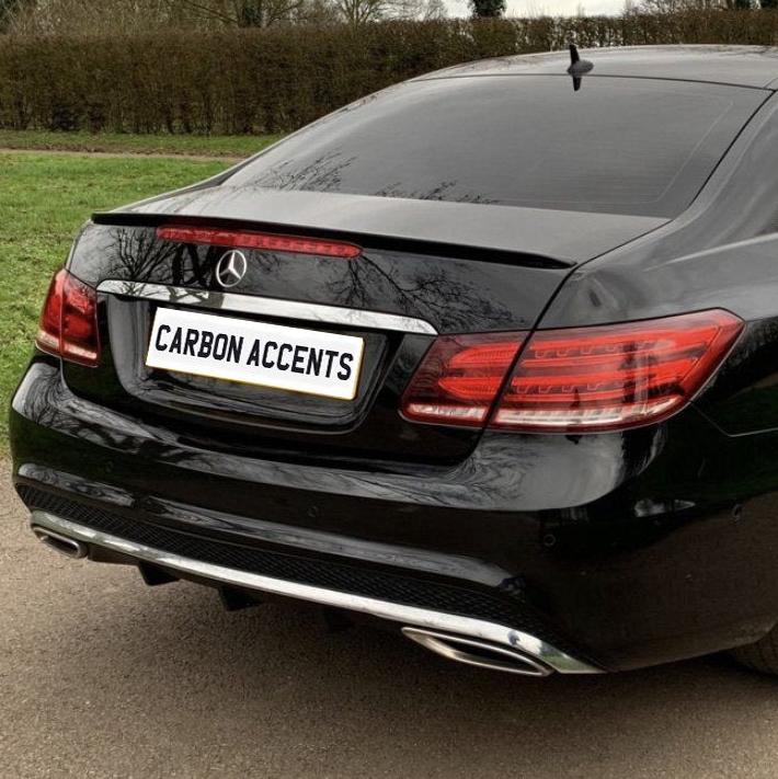 A black car with a "CARBON ACCENTS" license plate is parked on a paved road, surrounded by grass and trees in the background.