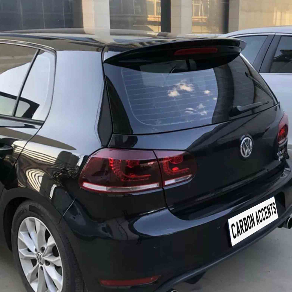 A black car is parked beside a white car in a lot. The rear view shows a Volkswagen logo and text reading "CARBON ACCENTS" on the bumper.