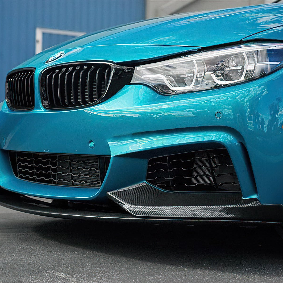 A blue car's front with a focus on the grille, headlight, and bumper detail, parked on a street. A blue building wall is visible in the background.