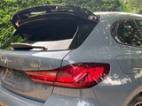 A gray BMW 118i hatchback with an open trunk is parked on a gravel driveway, surrounded by greenery, under a partly cloudy sky.