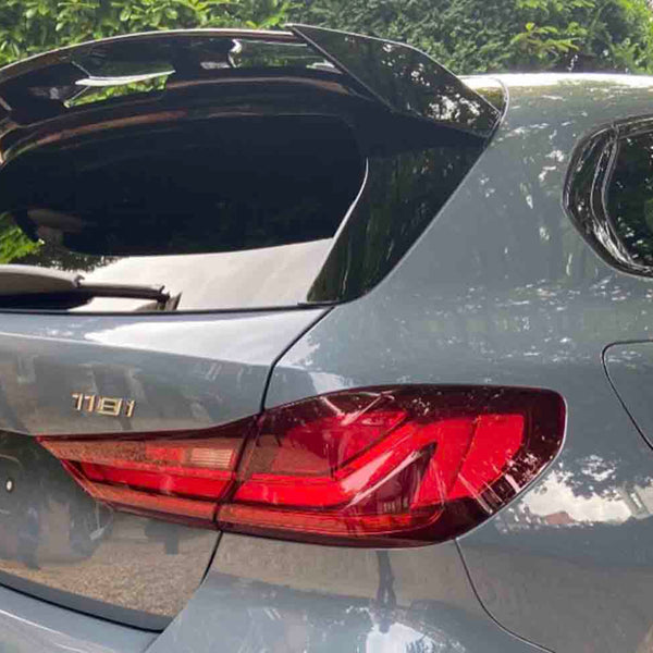 A gray BMW 118i hatchback with an open trunk is parked on a gravel driveway, surrounded by greenery, under a partly cloudy sky.