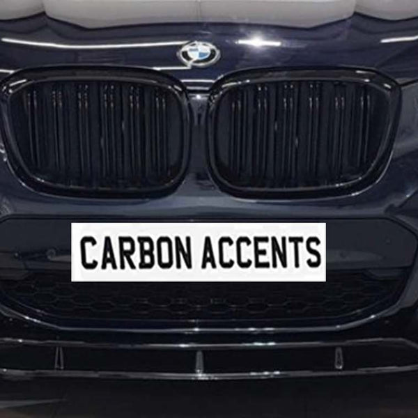 Car front grille with illuminated headlights; BMW logo above, and "CARBON ACCENTS" text on a simulated plate. The car is in a showroom setting with a tiled floor.