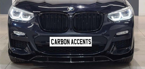 Car front grille with illuminated headlights; BMW logo above, and "CARBON ACCENTS" text on a simulated plate. The car is in a showroom setting with a tiled floor.