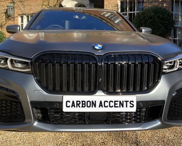 A BMW car parked on a gravel driveway features a prominent grille. The number plate reads "CARBON ACCENTS." A brick building with greenery is in the background.