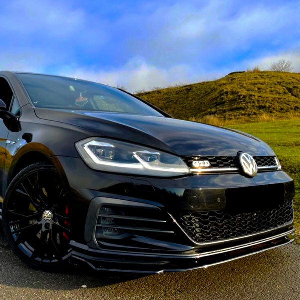 A black Volkswagen GTD car is parked on a paved surface, with headlights on, against a backdrop of grassy hills and a partly cloudy sky.