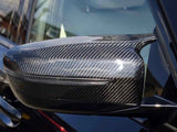 A carbon fiber car side mirror, reflecting light, is attached to the sleek black exterior of a vehicle, with a blurred urban background visible.